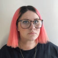 An image of Emily Sara, a white femme with short neon orange-ish pink hair. She is wearing large tortoiseshell glasses, a black t-shirt, two gold necklaces and has a faint scar across her neck. She is leaning up against a light grey wall with sunlight coming in from the right side of the frame.