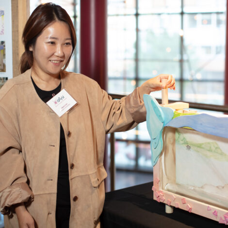 A woman holds the handle of a vertical roller at one side of a wooden tabletop mechanism. The roller spools paper across the display window of the mechanism; the paper is painted with delicate colors to show an outdoor scene.